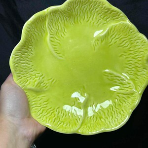 May include: A bright lime green ceramic plate with a glossy finish and a textured surface that mimics the appearance of a cabbage leaf. The plate features a scalloped, irregular edge and raised, vein-like details radiating from the center.