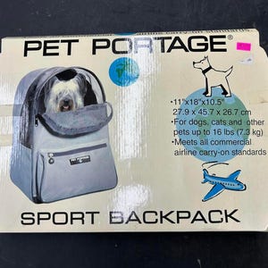 May include: A Pet Portage sport backpack in a light gray color with a mesh window showing a dog inside. The packaging box displays the text Pet Portage, dimensions of 11 inches by 18 inches by 10.5 inches, and states it is for pets up to 16 lbs.