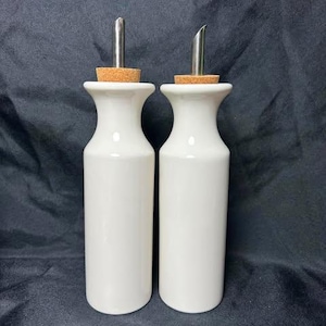 May include: Two white ceramic oil and vinegar dispensers with smooth, cylindrical bodies and flared necks. Each bottle is topped with a natural cork stopper and a silver-colored stainless steel pour spout for controlled liquid dispensing.