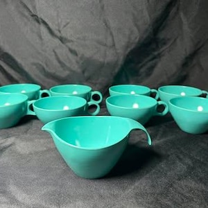 May include: A collection of eight matching turquoise cups, each featuring a smooth, rounded bowl shape and a single side handle. The set includes seven identical cups and one slightly larger, deeper cup with a distinct pouring spout.