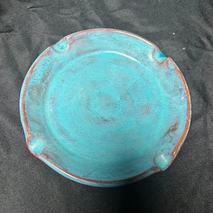 May include: A round ceramic ashtray featuring a vibrant turquoise blue glaze with visible reddish-brown undertones along the rim and textured surface. The piece includes three evenly spaced notches around the edge for resting items securely.