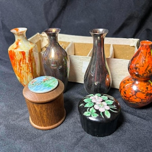 May include: A collection of four small vases and two lidded containers. Two vases are gourd-shaped with orange and red mottled patterns, while two are metallic. One wooden container has a circular lid with a blue landscape, and one black container features a pink floral design.
