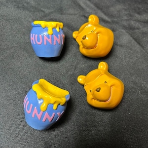 May include: Two blue ceramic honey pots with yellow drips and the word HUNNY in pink letters, paired with two yellow bear head figurines. The bear heads feature small ears, round noses, and subtle smiles, while the pots have a rounded shape.