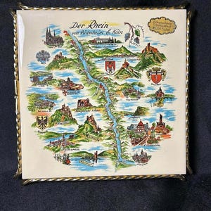 May include: A square ceramic tile featuring a colorful illustrated map of the Rhine River from Rudesheim to Koln, titled Der Rhein von Rudesheim b. Koln. The tile is framed in a twisted gold-tone metal border and displays various landmarks.
