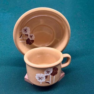 May include: A light tan ceramic cup and matching saucer set. Both pieces feature a decorative floral pattern consisting of white and dark brown flowers with thin stems. The cup has a rounded handle on the side and a smooth, glossy finish throughout.