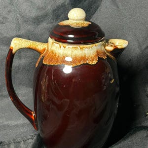 May include: A ceramic teapot featuring a deep brown glossy glaze on the body and a contrasting cream-colored drip glaze around the rim, handle, and lid. The lid has a small, rounded knob, and the handle is curved, extending from the top to the side.