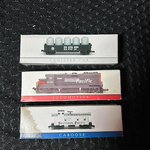 May include: Three model train cars in individual white boxes. The top box shows a black Southern Pacific canister car. The middle box displays a dark gray Southern Pacific locomotive numbered 9725. The bottom box features a white Southern Pacific caboose.