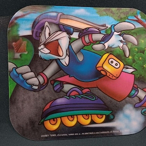 May include: A rectangular graphic featuring Bugs Bunny wearing a purple helmet, blue tank top, and pink shorts while rollerblading on a path. The character wears blue wrist guards and knee pads, with a yellow pouch at the waist and orange wheels.