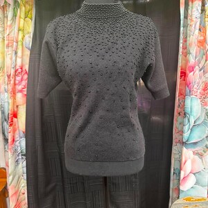May include: A black short-sleeved sweater featuring a high mock neck collar. The garment is adorned with numerous small, round black beads that are densely clustered around the neckline and become more sparse toward the hem of the sweater.