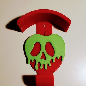 Può includere: Un supporto da parete rosso stampato in 3D a forma di mela avvelenata con una mela verde e un design a forma di teschio. Il supporto ha una parte superiore curva per appendere oggetti.