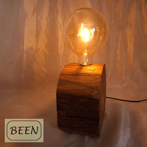 Könnte beinhalten: Eine Tischleuchte mit einem dicken, zylindrischen Stück aus natürlichem braunem Holz als Basis, gekrönt von einer großen, klaren, runden Glasglühbirne, die ein warmes, orangefarbenes Licht ausstrahlt. Das Wort BEEN ist schwarz auf einem hellbraunen Rechteck gedruckt.