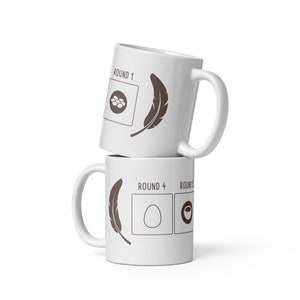 May include: Two white ceramic mugs stacked on top of each other. The top mug has the text "ROUND 1" and a feather design. The bottom mug has the text "ROUND 4" and a feather design. Both mugs have a brown design.