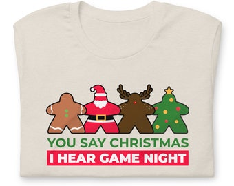 Puede incluir: Una camiseta beige con un gráfico de cuatro hombres de jengibre que representan personajes navideños: un hombre de jengibre, Santa Claus, un reno y un árbol de Navidad. El texto "You say Christmas I hear game night" está impreso debajo del gráfico.