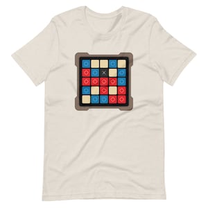May include: Beige t-shirt featuring a colorful graphic of a game board. The board has a grid of squares in red, blue, and yellow, with an 'X' in the center. The game board is framed in a brown border.