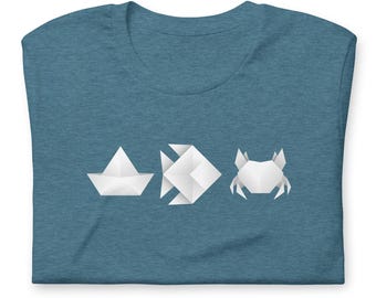 May include: A blue t-shirt featuring three white origami figures: a boat, a fish, and a crab. The origami designs are simple and geometric, creating a minimalist aesthetic. The shirt is made of a soft material.