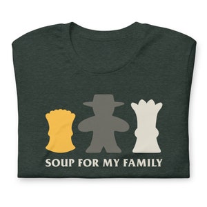 Könnte beinhalten: Dunkel meliertes grünes T-Shirt mit einer Grafik von drei stilisierten Spielsteinen in Gelb, Grau und Weiß. Der Text "SOUP FOR MY FAMILY" ist darunter in Weiß gedruckt. Das T-Shirt ist gefaltet.