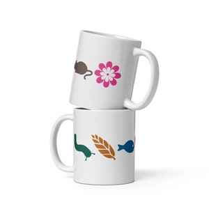 Könnte beinhalten: Zwei weiße Keramikbecher, die übereinander gestapelt sind. Der obere Becher hat eine braune Maus, eine rosa Blume und einen weißen Hintergrund. Der untere Becher hat eine grüne Schnecke, eine braune Weizenähre und einen blauen Fisch auf weißem Hintergrund.