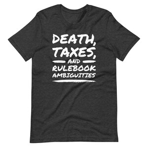 May include: Dark gray t-shirt with white text that reads "DEATH, TAXES, AND RULEBOOK AMBIGUITIES". The text is in a bold, handwritten style. The shirt is a short-sleeved crew neck.