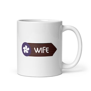 May include: White ceramic mug with a brown wooden sign that says "WIFE" with a purple flower icon.