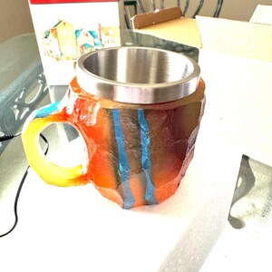 May include: A colorful, rock-shaped mug with a stainless steel interior. The mug has a yellow handle and blue stripes.