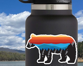 May include: A black bear silhouette sticker with a sunset and mountain landscape design. The sticker is 3 inches by 2 inches and features a retro color palette of orange, yellow, blue, and gray. The sticker is on a black water bottle with the text "Life Outside Sticker Company".