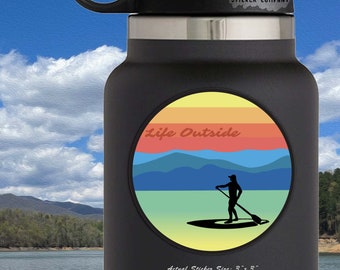 May include: A black water bottle with a round sticker featuring a sunset scene with mountains and a person paddleboarding. The sticker says "Life Outside" in a retro font. The sticker is 3 inches by 3 inches.