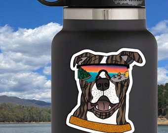 May include: A sticker of a brindle dog wearing sunglasses with a sunset and mountains reflected in the lenses. The sticker is on a black water bottle. The text "Life Outside Sticker Company" is on the bottle. The sticker is 2.5 inches by 2 inches.