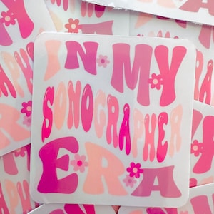 May include: Pink and white sticker with the text "In My Sonographer Era" in a retro font. The sticker is decorated with small pink flowers.