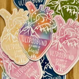 Puede incluir: Una colección de pegatinas en forma de corazón con el texto "ULTRASOUND is a work of heart". Las pegatinas presentan diseños de montañas y árboles, con colores como azul, rosa, amarillo, verde y degradados arcoíris.