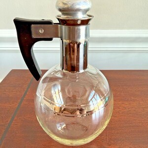 May include: A clear glass decanter with a round body, a silver-colored metal band, and a black handle. The stopper is silver-colored. The decanter sits on a brown surface.