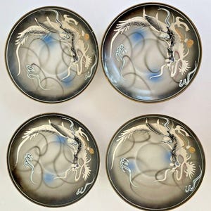 May include: Set of four decorative plates with a dragon design. Each plate features a black and gray gradient background with a white and gold dragon illustration. The plates have a gold rim and a blue accent.