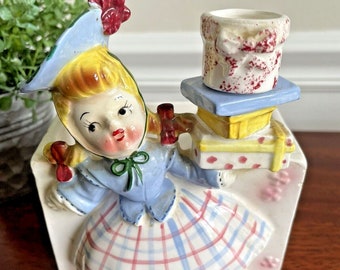 May include: A vintage ceramic planter shaped like a girl in a blue dress and red hat, holding a stack of presents. The planter has a white background with pink and blue plaid on the skirt.