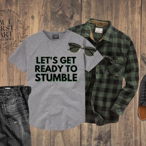May include: A grey t-shirt with the text "Let's get ready to stumble" in green letters. The t-shirt is paired with black jeans, a green and black plaid shirt, and black sneakers.