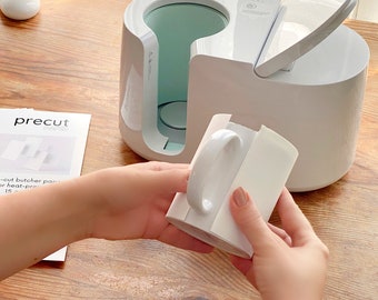May include: White mug press machine with a white mug being inserted into the machine. The mug press machine has a white lid that is open. The mug is being held by a person's hands. The machine has a digital display with the text "Precut" on it.