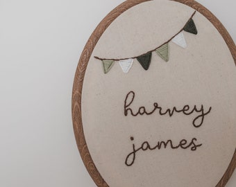 May include: A brown wooden embroidery hoop with white fabric inside. The fabric has green and white triangle flags stitched on it and the names "harvey james" embroidered in brown thread.