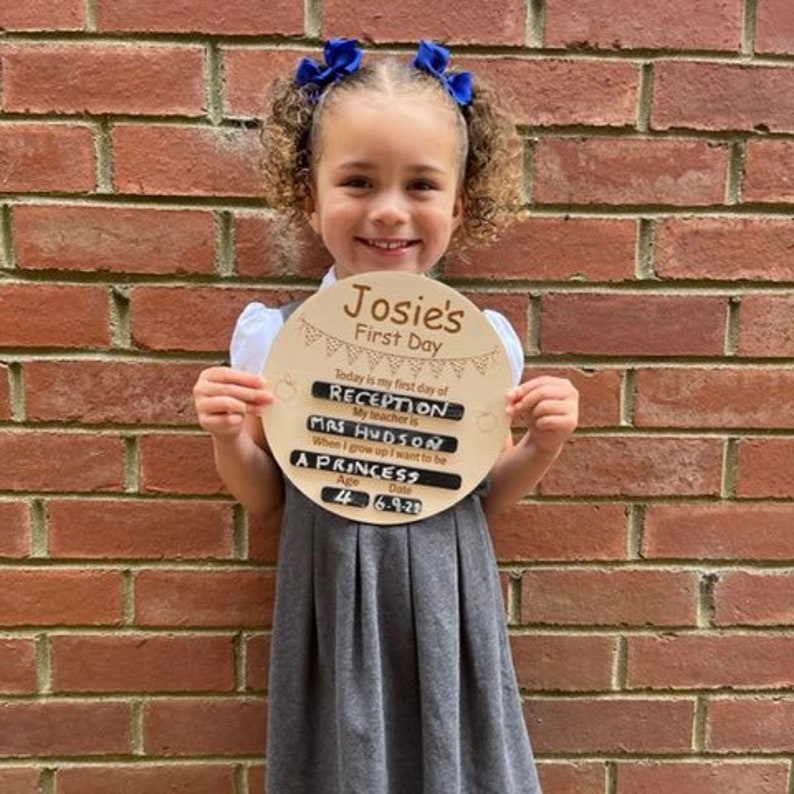 Pu&ograve; includere: Un cartello di legno con il testo "Josie's First Day" e uno striscione di bandierine. Il cartello include anche il testo "Today is my first day of Reception, My teacher is Mrs Hudson, When I grow up I want to be A Princess, Age 4, Date 6-9-21".