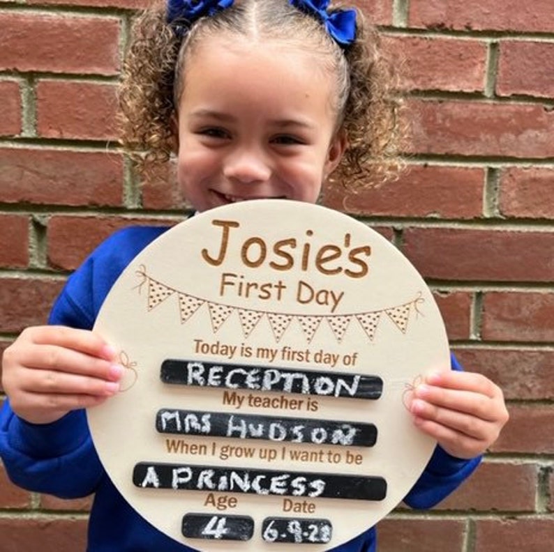 Pu&ograve; includere: Un cartello rotondo di legno con il testo "Josie's First Day" e uno striscione di bandiere. Il cartello ha spazi vuoti per scrivere con il gesso. Il testo "Today is my first day of Reception. My teacher is Mrs Hudson. When I grow up I want to be a Princess. Age 4. Date 6-9-22" &egrave; scritto sul cartello.