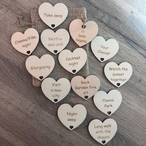 May include: A set of 12 wooden heart-shaped date night ideas. The hearts are in a hessian bag and feature phrases like "Take away", "Netflix and chill", "12 Date Nights", "Stargazing", "Cocktail night", "Your Choice!", "Visit a new city", "Back Garden fire pit", "Watch the sunset together", "Theme Park", "Night away", and "Long walk with the doggos".