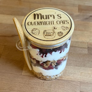 May include: A glass jar containing layered overnight oats with a wooden lid. The lid is engraved with "Mum's OVERNIGHT OATS" and food icons. A wooden spoon is attached. The jar contains layers of yogurt, granola, and berries.
