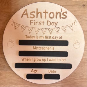 Pu&ograve; includere: Un cartello rotondo in legno con il testo "Ashton's First Day" e uno stendardo di bandierine con pois. Il cartello ha spazi vuoti per scrivere nel testo "Today is my first day of", "My teacher is", "When I grow up I want to be", "Age", e "Date".