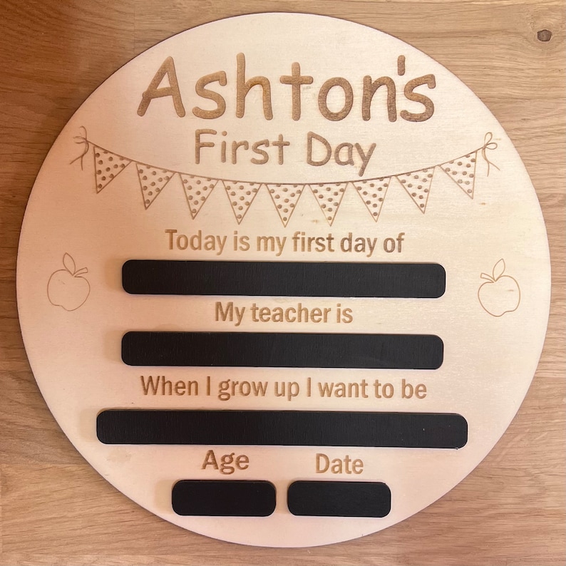 Pu&ograve; includere: Un cartello rotondo in legno con il testo "Ashton's First Day" e uno stendardo di bandierine con pois. Il cartello ha spazi vuoti per scrivere nel testo "Today is my first day of", "My teacher is", "When I grow up I want to be", "Age", e "Date".
