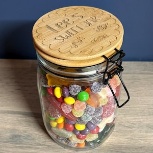 May include: A clear glass jar filled with assorted colourful sweets, featuring a bamboo lid with a scalloped edge and a black metal wire clamp closure. The lid is laser-engraved with the text Lee's Sweet Jar and small sweet illustrations.