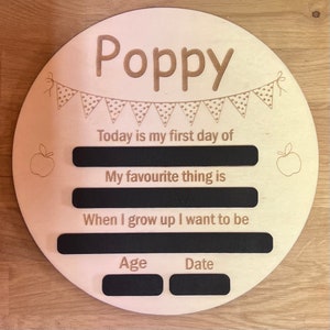 Pu&ograve; includere: Un cartello rotondo in legno con il nome "Poppy" in alto. Il cartello ha uno striscione con i pois e uno spazio per scrivere "Oggi &egrave; il mio primo giorno di", "La mia cosa preferita &egrave;", "Quando crescer&ograve; voglio essere", "Et&agrave;", e "Data".
