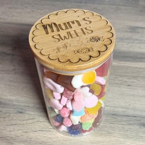 May include: A glass jar filled with a selection of colourful sweets, topped with a wooden lid. The lid is engraved with the words "Mum's SWEETS" and candy illustrations. The jar contains a variety of sweets in different shapes and colours.