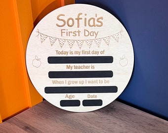 May include: A round wooden sign with the text "Sofia's First Day" and spaces for writing the child's first day of school information. The sign has a black chalkboard background and a decorative banner with polka dots.