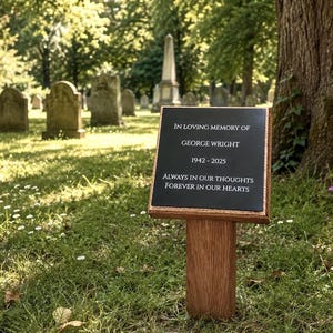 May include: A rectangular black memorial plaque mounted on a wooden stake. The plaque features white engraved text reading: IN LOVING MEMORY OF GEORGE WRIGHT 1942-2025 ALWAYS IN OUR THOUGHTS FOREVER IN OUR HEARTS. The wooden stake is set into the ground.
