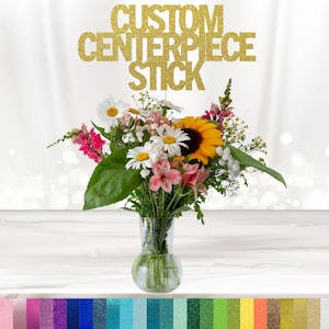 May include: Gold glitter "CUSTOM CENTERPIECE STICK" sign with a bouquet of flowers in a clear glass vase. The flowers include white daisies, pink flowers, and a yellow sunflower.