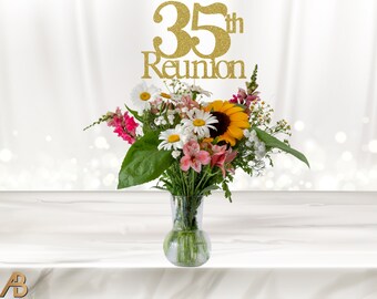 May include: A gold glitter 35th Reunion cake topper with the words "35th Reunion" on it. The topper is placed on top of a bouquet of flowers in a clear glass vase.