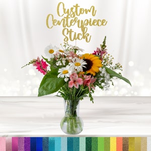 May include: Gold glitter "Custom Centerpiece Stick" sign with a bouquet of flowers in a clear glass vase. The flowers include daisies, sunflowers, and pink flowers. The vase is on a white wooden surface.