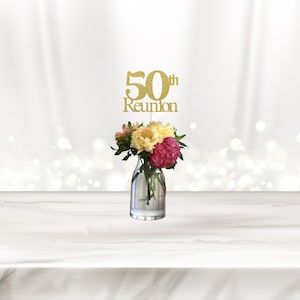 May include: Gold glitter 50th reunion centerpiece with the words "50th Reunion" on a stick. The stick is inserted into a clear glass vase with pink, yellow, and white flowers.